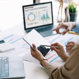 Two people conferring over financial reports on laptop, calculator and tablet.