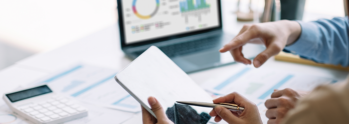 Two people conferring over financial reports on laptop, calculator and tablet.