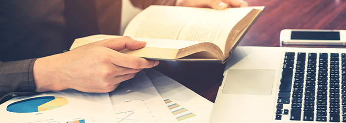 Person holding a financial book in their heads with their laptop above and financial reports on the table