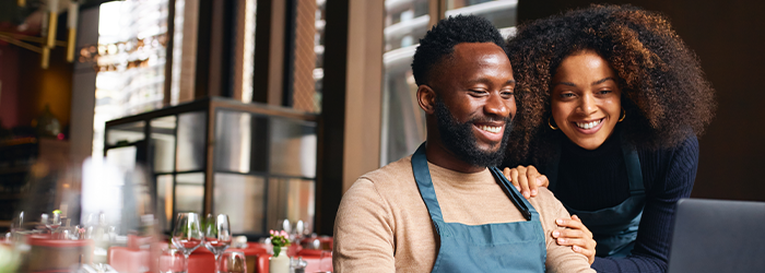 Man with apron seated and smiling at laptop while holding credit card, woman leaning in and smiling as well