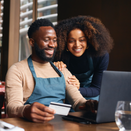 Man with apron seated and smiling at laptop while holding credit card, woman leaning in and smiling as well