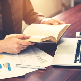 Person holding a financial book in their heads with their laptop above and financial reports on the table